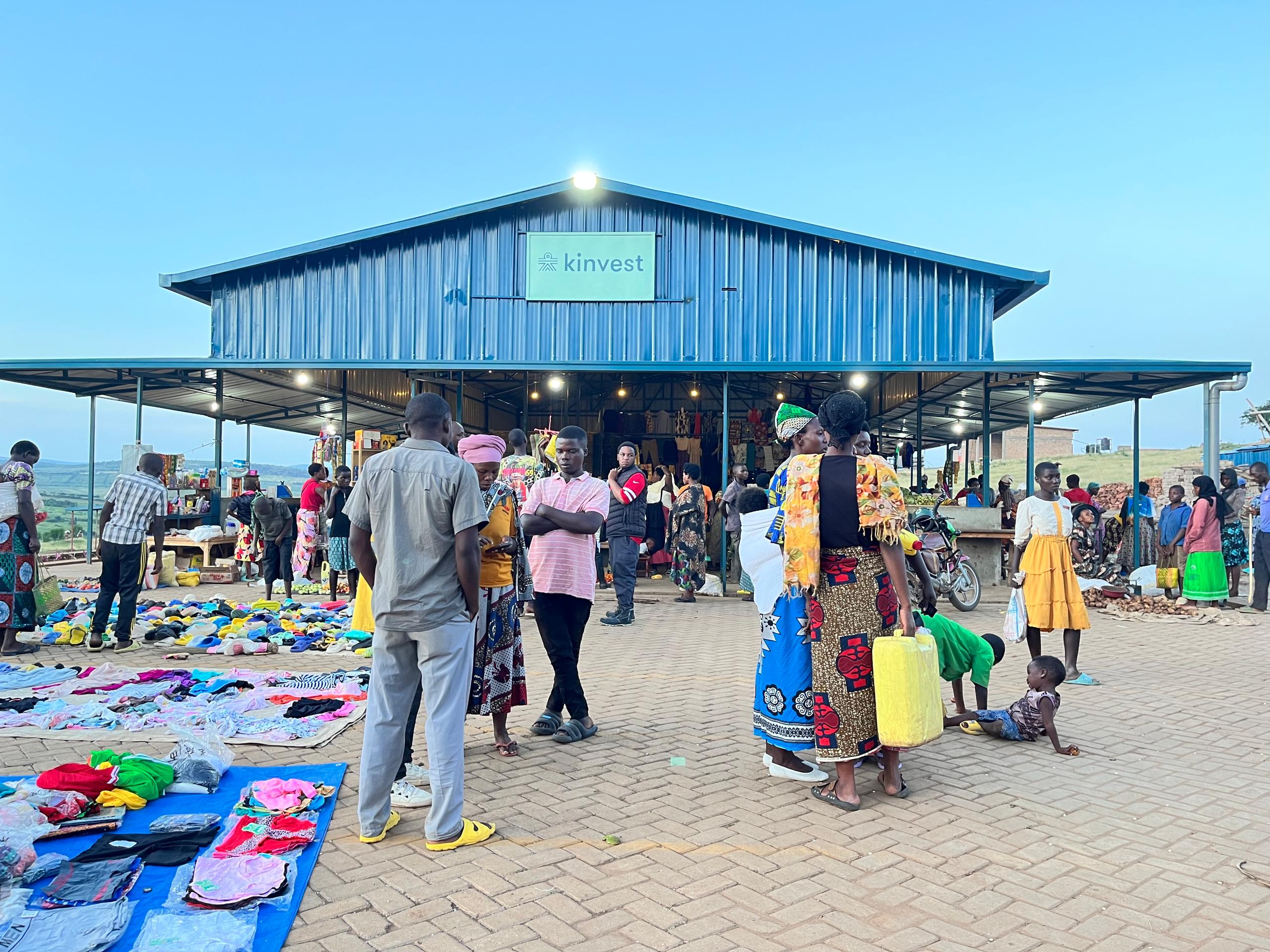 A marketplace full of people in rural Rwanda. The blue covered market place is in the background while in the foreground people interact with market stalls laid out on a concrete plaza.